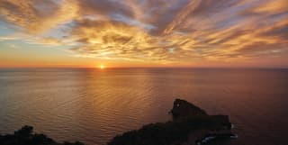 Sun melts on the horizon in a glorious orange sunset display that tints the sea around Na Foradada's famous holed rock.