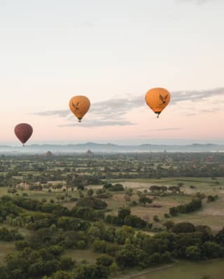 View over Myanmar's mountain-fringed fields as three hot air balloons in reds and yellows glide through a pale peach sky.